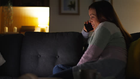 Primer-Plano-De-Una-Mujer-Sonriente-Sentada-En-Un-Sofá-En-Casa-Por-La-Noche-Hablando-Por-Teléfono-Móvil-2